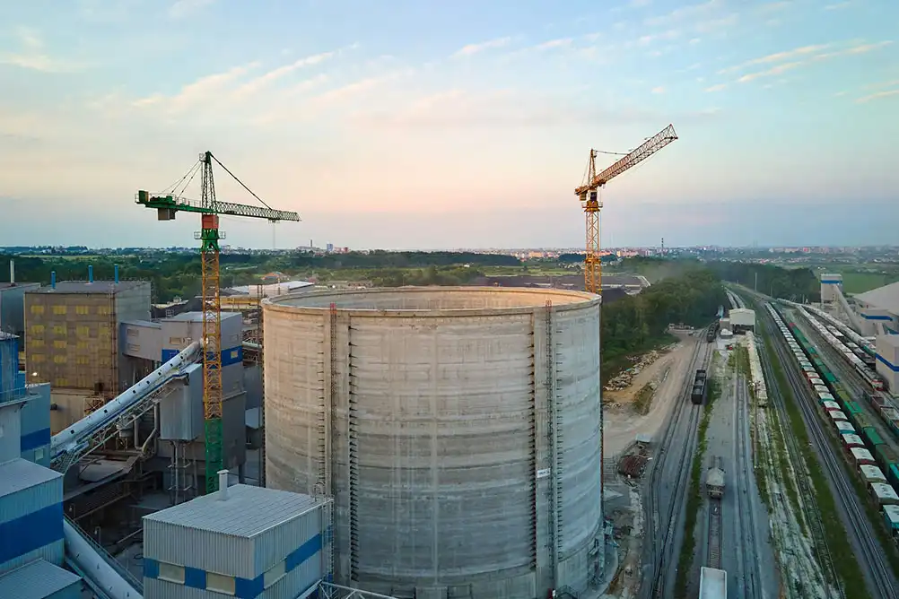 A large industrial construction site featuring a massive cylindrical concrete storage tank surrounded by cranes and factory buildings